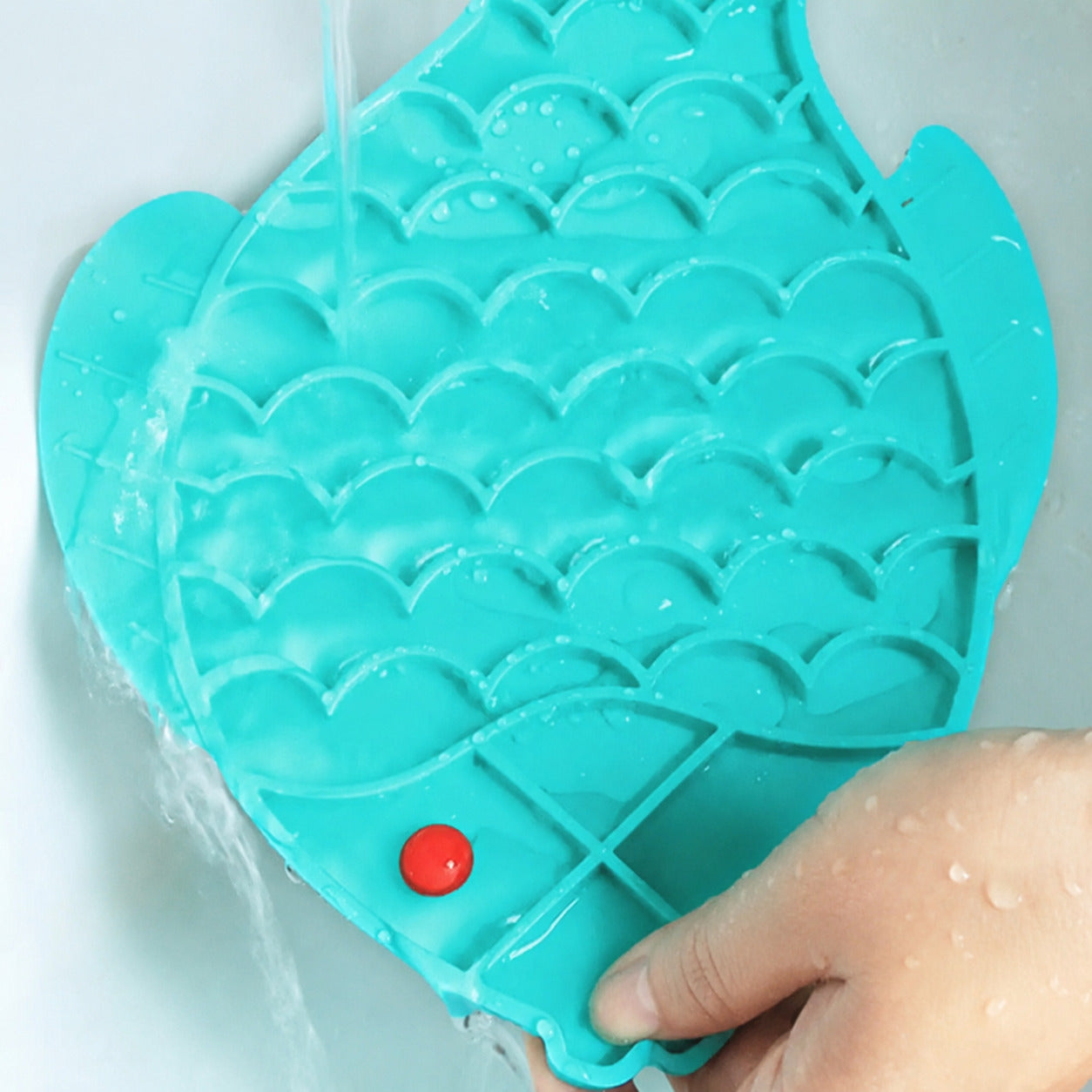 Blue fish-shaped slow feeder lick mat being rinsed clean under running water